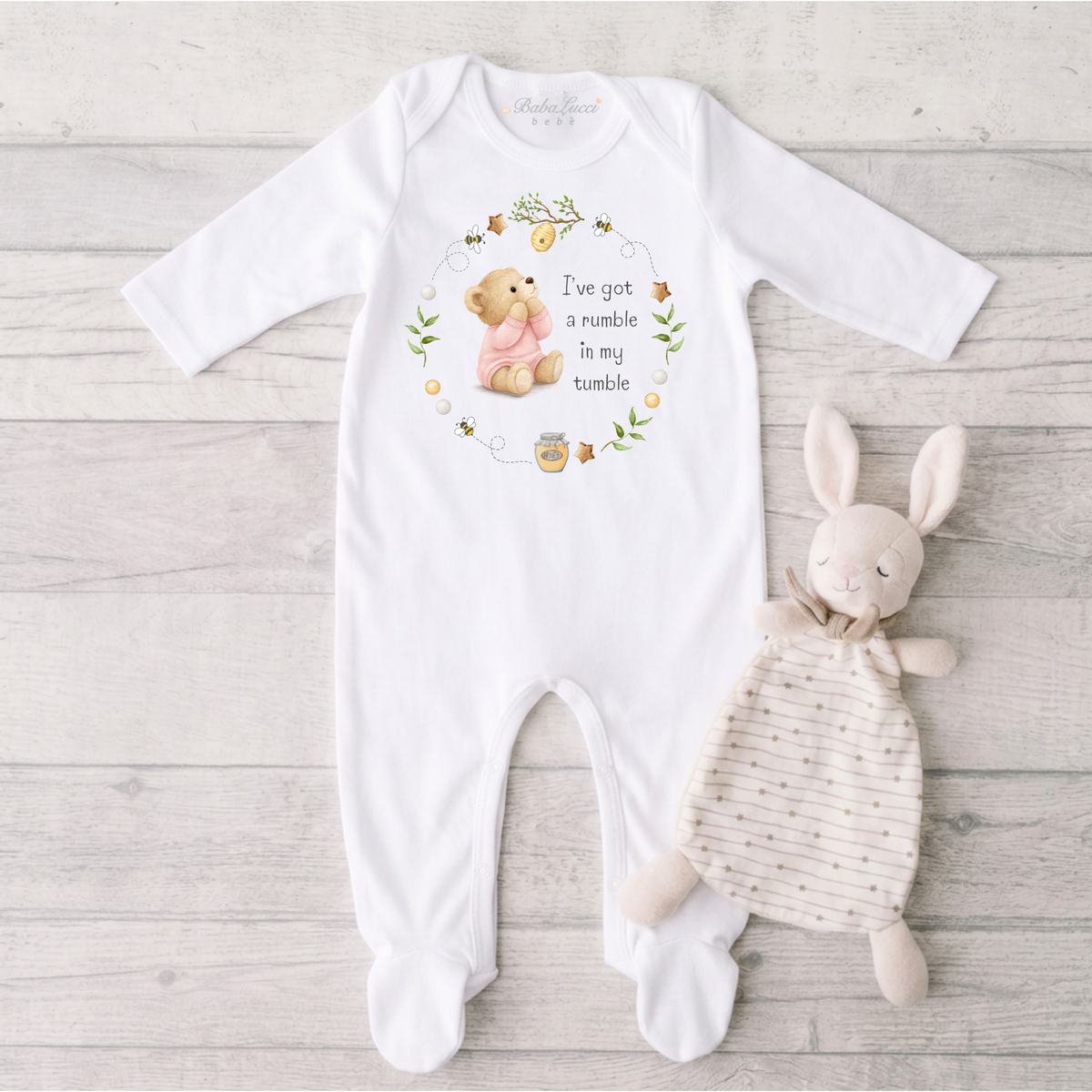 A white long-sleeve baby sleepsuit, featuring a printed graphic of a teddy bear in a wreath with the text 'I've got a rumble in my tumble.' 