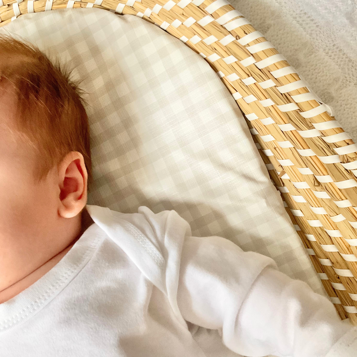 A close-up lifestyle shot of a baby lying in a woven wicker bassinet, wearing a white long-sleeve Sleepsuit. The Close-up shot is to show the envelope neckline detail of the garment.
