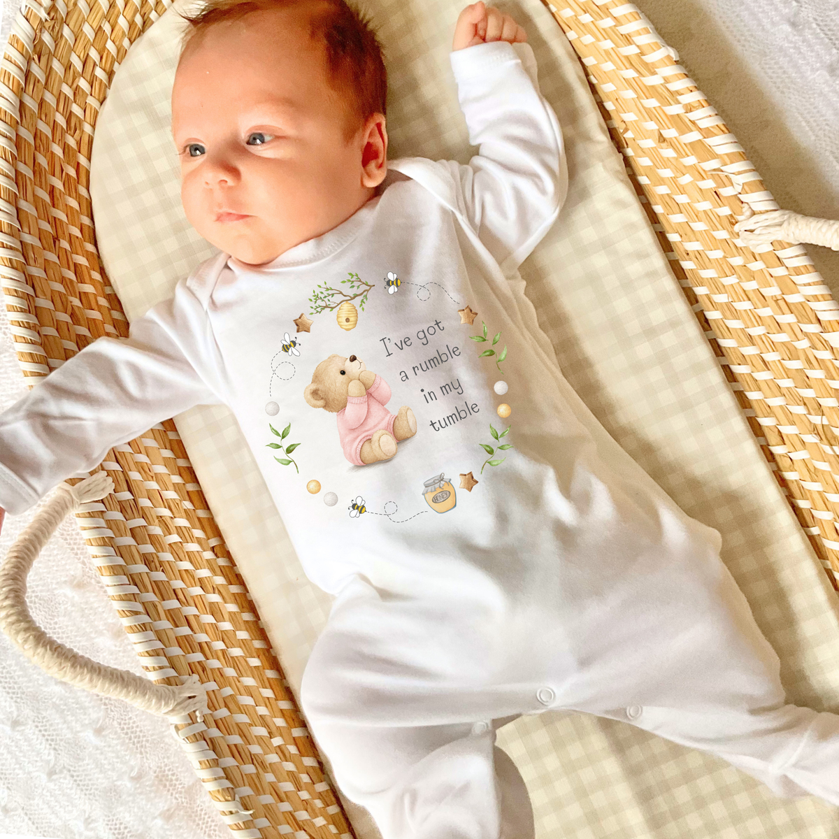 A baby wearing a white long-sleeve footed sleepsuit with a teddy bear graphic and the text 'I've got a rumbly in my tumble'.